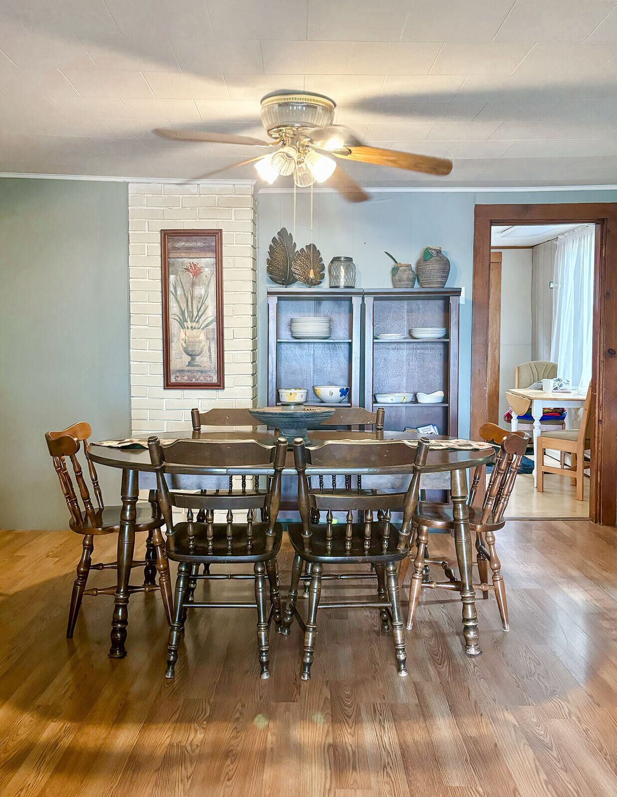 A dining room with a round wooden table and six chairs, a ceiling fan, and a brick-patterned wall with a framed picture.