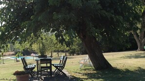Un espace extérieur aménagé avec une table et des chaises sous un arbre, une piscine en arrière-plan et une pelouse verdoyante.