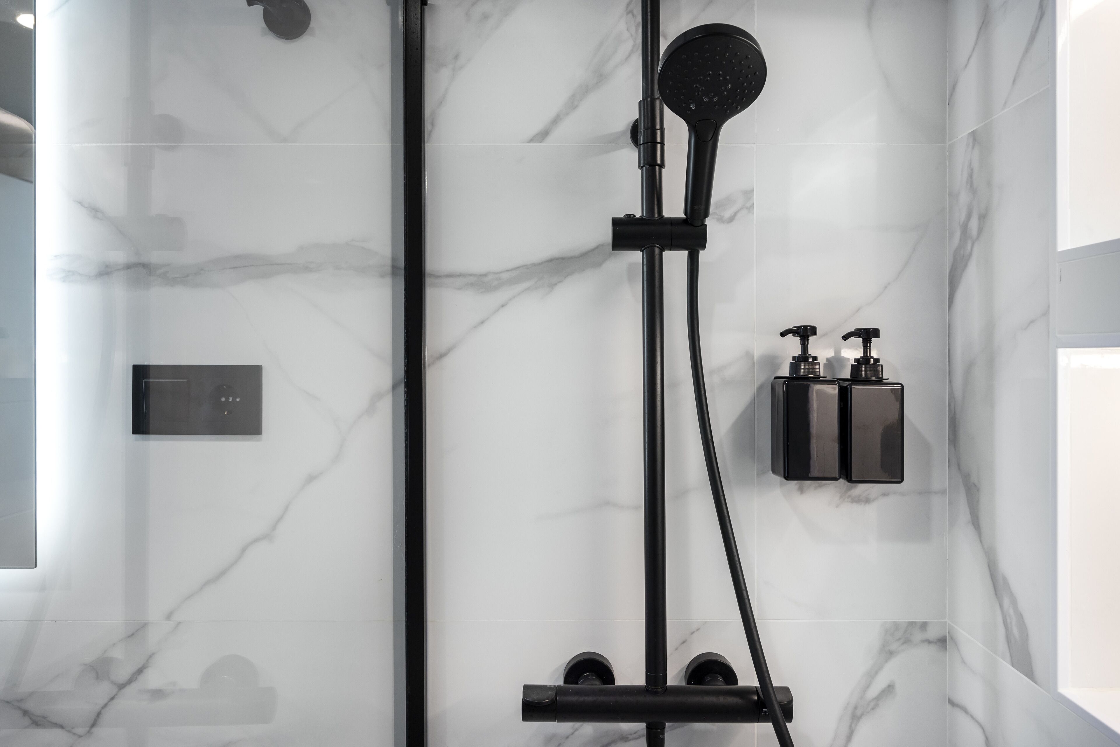 Ein modernes Badezimmer mit Marmorwand, Brausekopf in Schwarz und zwei schwarzen Seifenspendern an der Wand.