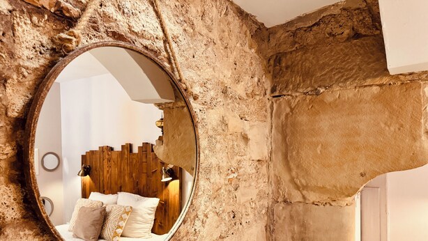A circular mirror embedded in a textured stone wall, reflecting a bed with a wooden headboard and pillows. - La Chaumière - BedinShop - historic center - France (Romans-sur-Isère)
