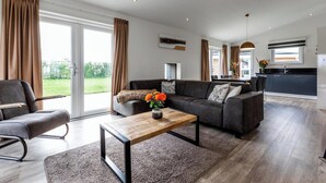 A modern living room with a sofa, a coffee table, and a view of the garden through large sliding doors. - Schaelsbergerbosch - Fireplace & Sauna (Bemelen)