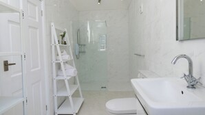 Une salle de bain moderne avec un lavabo blanc, des toilettes et une douche à parois de verre. On y trouve une étagère blanche avec des serviettes et une petite plante.