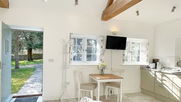 Une chambre avec un lit, un téléviseur fixé au mur, une petite table avec des chaises, et une vue sur le jardin par la porte ouverte.
