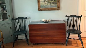 A room with a wooden cabinet, two chairs, a table with a bowl, and a framed painting on the wall.