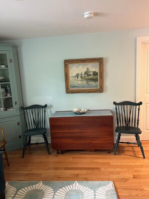 A room with a wooden cabinet, two chairs, a table with a bowl, and a framed painting on the wall.