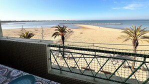 Vista a la playa desde un balcón, con el mar, palmeras y un muelle distante.