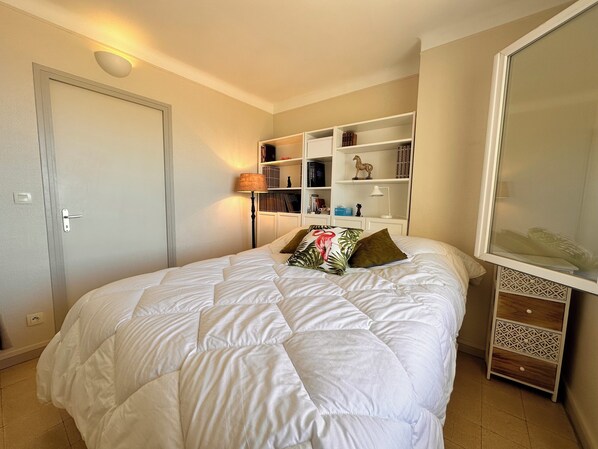Una cama bien tendida con edredón blanco y dos almohadas, una lámpara de noche, una estantería con libros y objetos decorativos, y una puerta con interruptor de luz.