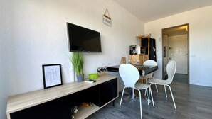 A modern living space with a flat-screen TV mounted on the wall, a dining area with white chairs, a wooden console table, and a kitchenette area.