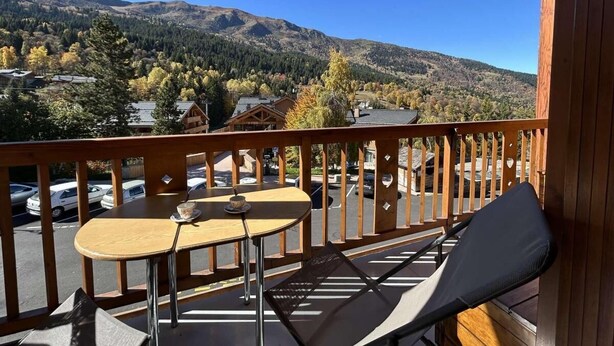 Een balkon met een houten hekwerk, twee tafels en een fauteuil met kussens.