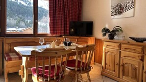 Een gezellige eethoek met een houten tafel, stoelen en een bank. Een televisie is aan de muur bevestigd, en er is een raam met gordijnen.