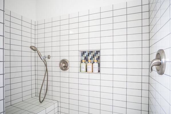En duschhörna med vita kakelväggar, ett duschmunstycke som kan hållas i handen och en liten hylla med tre flaskor schampo och balsam.