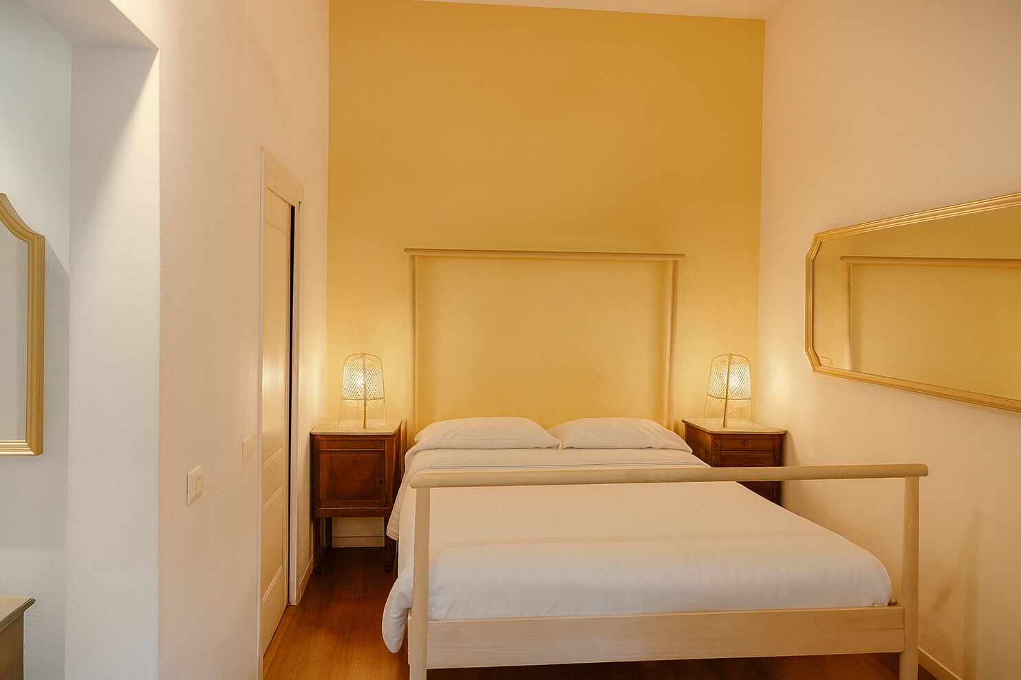 Quarto de hotel pequeno com uma cama de solteiro, mesas de cabeceira, um espelho e uma porta.