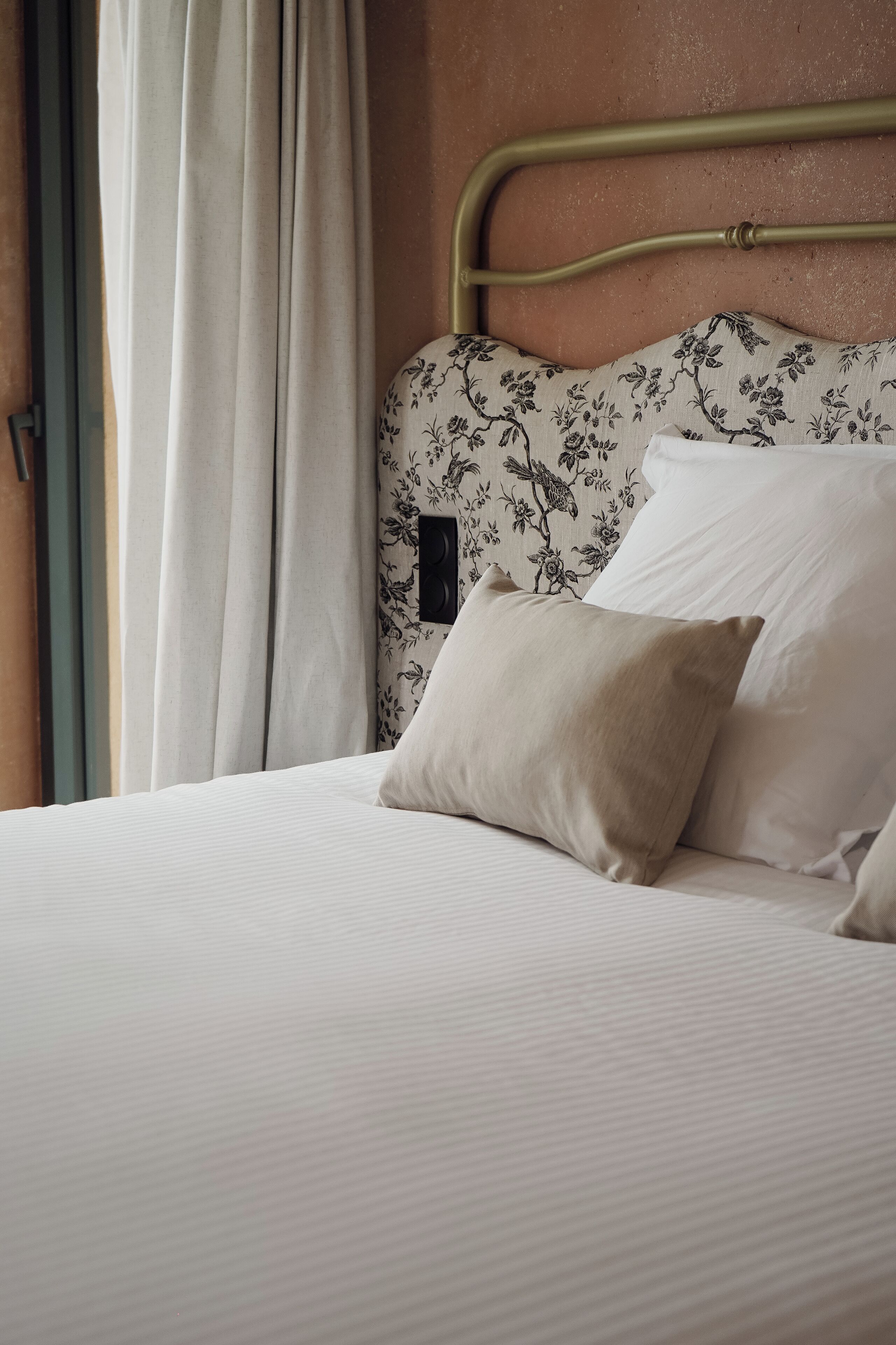 A neatly made bed with white linens and two beige pillows, a floral-patterned headboard, and a window with white curtains.