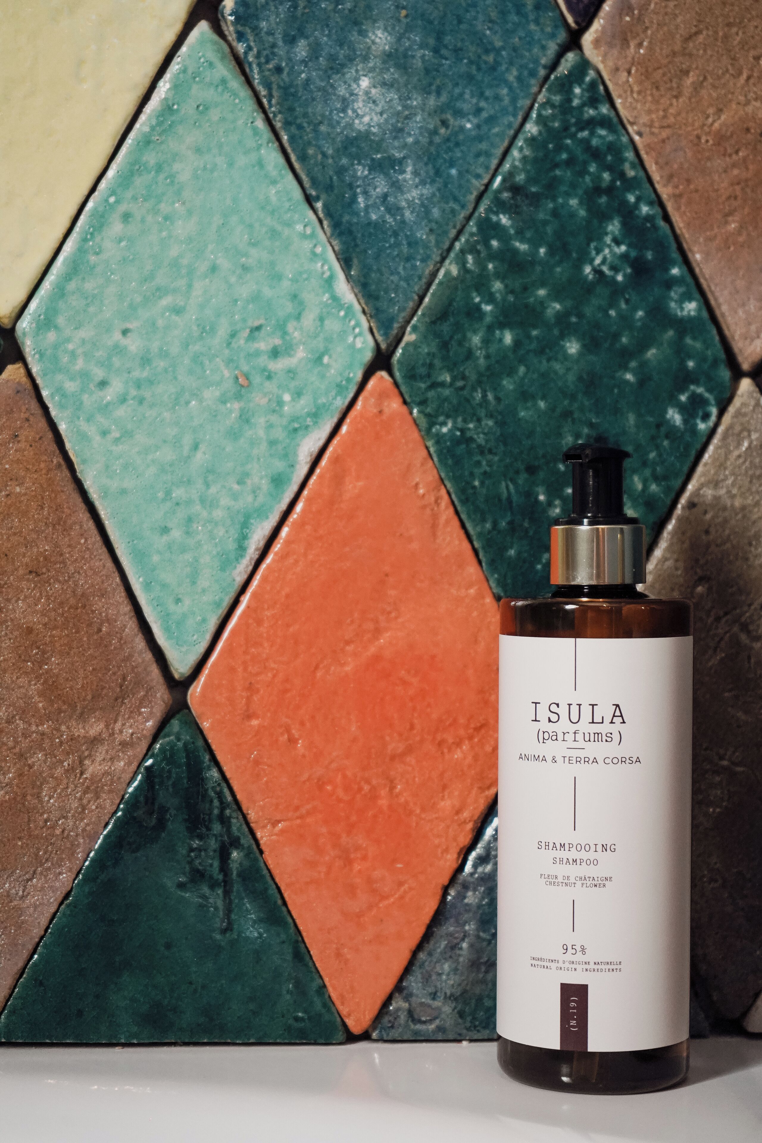 A bottle of Isula shampoo with a pump dispenser, set against a background of colorful, geometric tiles.