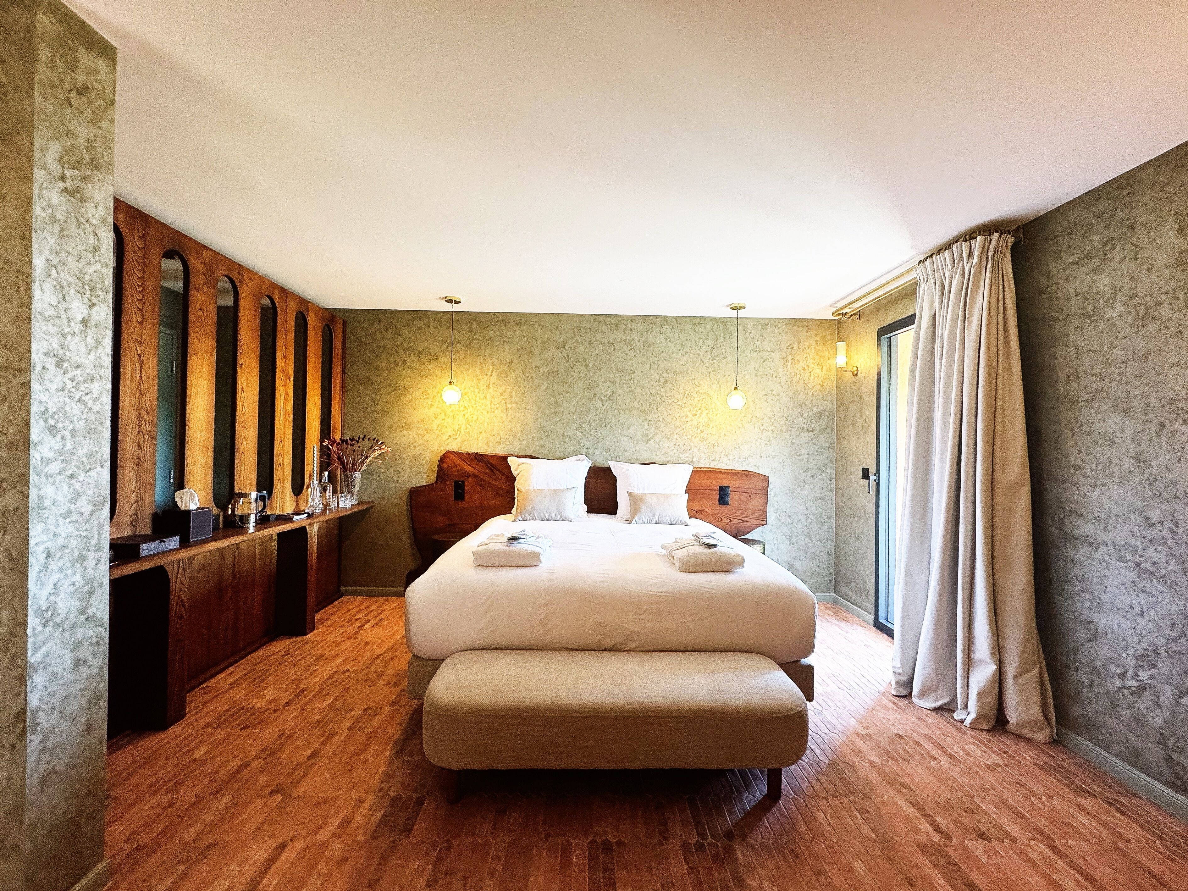 Habitación de hotel con una cama grande, cabecera de madera y un banco de madera. Hay una mesita con un florero y un espejo. El suelo es de madera y hay una ventana con cortinas.