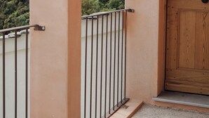 A balcony with a metal railing, a wooden door, and a view of a forested hillside. - DOMAINE SANTA MARGHERITA (Castellare-di-Casinca)