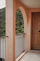 Un balcón con barandilla de metal, una puerta de madera y vistas a una ladera boscosa.