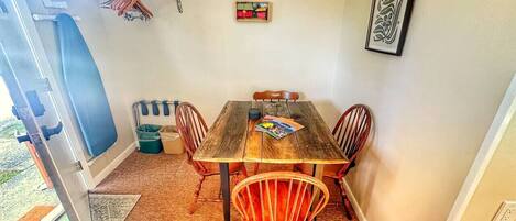Uma sala de jantar com mesa de madeira e quatro cadeiras, uma pequena prateleira com toalhas e um quadro na parede.