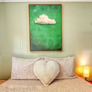 Ein Schlafzimmer mit einem Bett, einem herzförmigen Kissen und einem gerahmten Gemälde einer Wolke an der Wand.