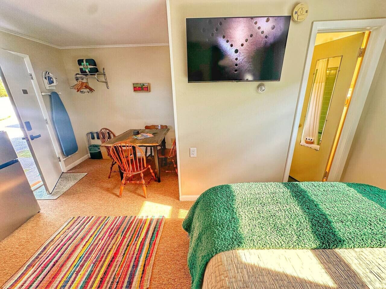 A compact room with a bed, a TV mounted on the wall, a dining area with a table and chairs, and a small kitchenette.
