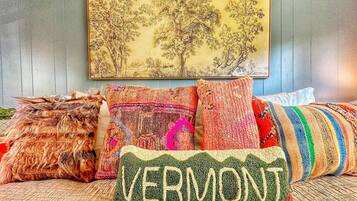 A bed with multiple colorful pillows, including one with "VERMONT" embroidered on it, and a painting of trees on the wall behind.