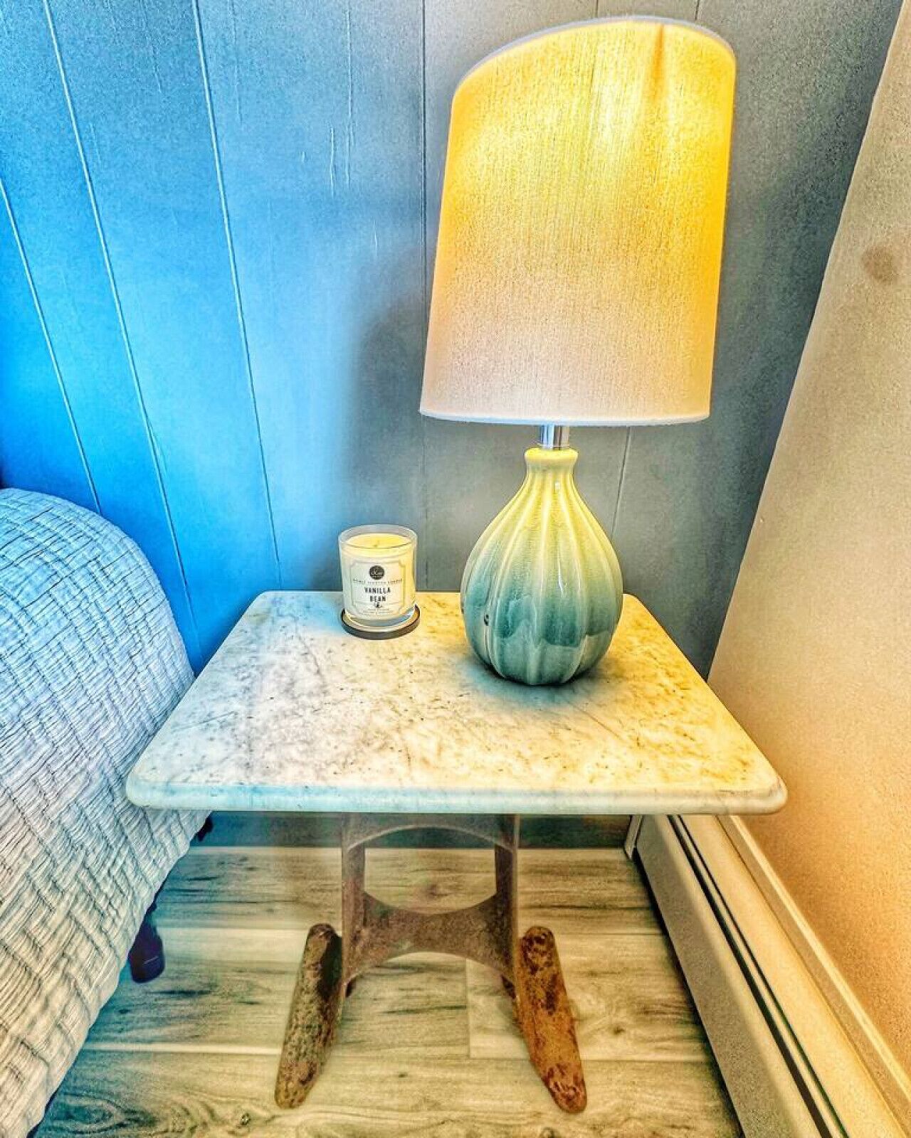 A small bedside table with a lamp, a candle, and a container of vanilla bean candles.