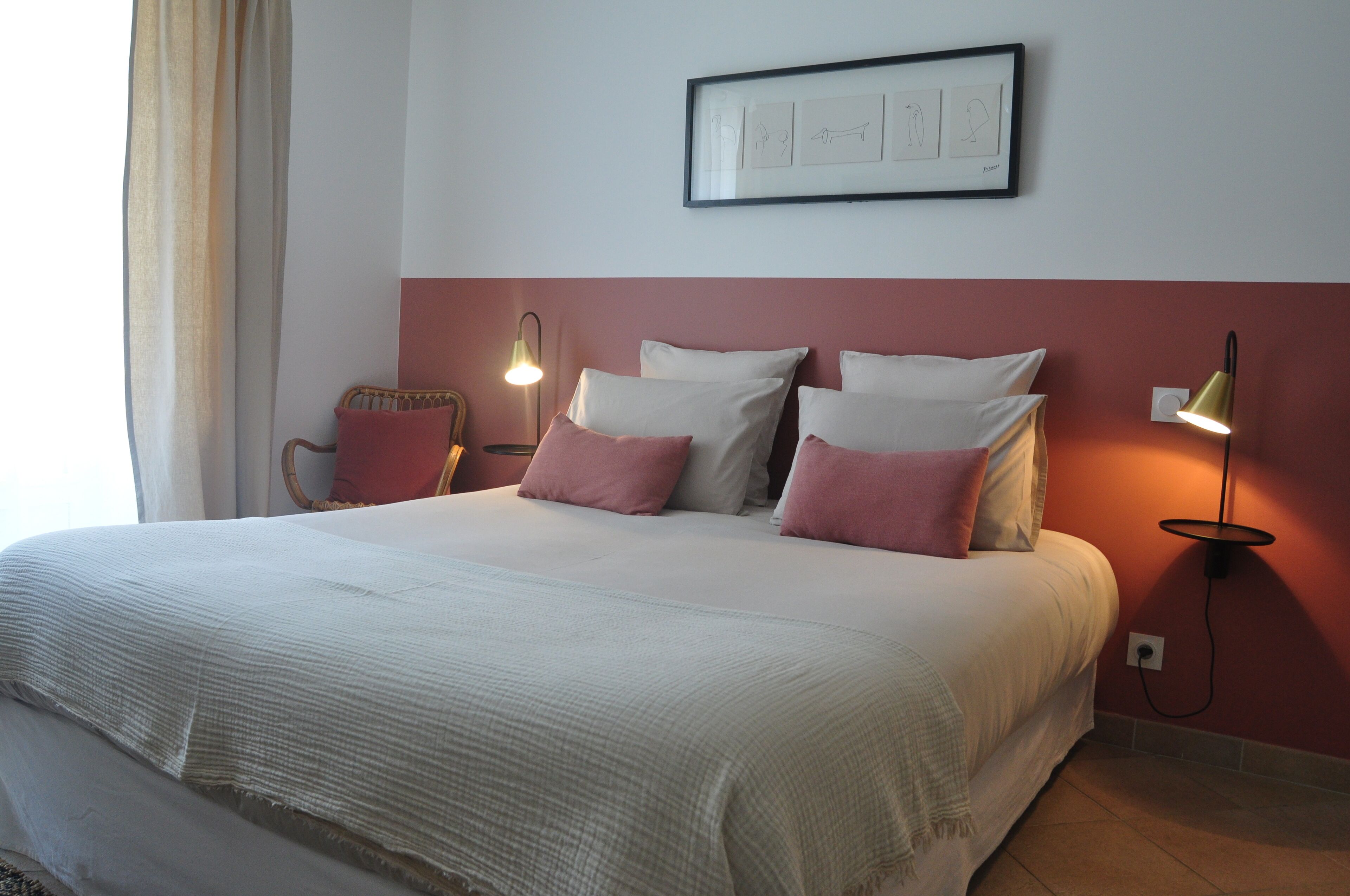 Una cama bien hecha con ropa de cama blanca y rosa, una obra de arte enmarcada encima y dos lámparas fijadas a la pared.