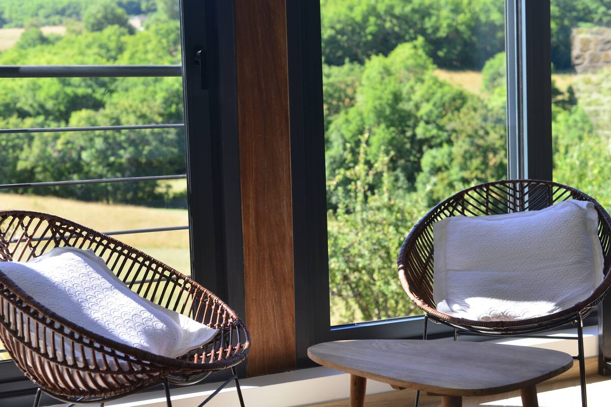 Zwei Korbstühle mit Kissen, ein kleiner Holztisch und ein großes Fenster mit Blick auf Grünflächen.