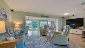 Ein großzügiges Wohnzimmer mit einer Couch, Sesseln, einem Couchtisch und einem Fernseher. Eine Tür führt zu einem Balkon mit Blick ins Freie.