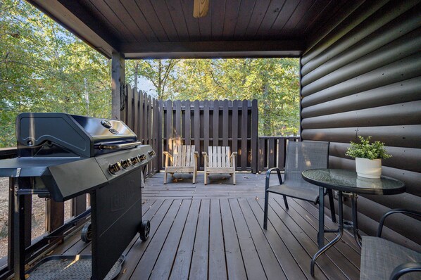A covered deck with a grill, two Adirondack chairs, a table with a potted plant, and a wooden fence. - Jax Ridge-Studio 1 Bedroom (Broken Bow)