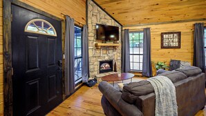 A cozy cabin interior with a stone fireplace, a large flat-screen TV mounted above it, a comfortable sofa, and a dining table. - Jax Ridge-Studio 1 Bedroom (Broken Bow)