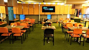 A hotel dining area with tables and orange chairs, a TV screen displaying 'CLASSY STAY', and a green carpeted floor. - Hotel Bramha inn (Pune)
