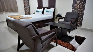 A bedroom with a bed, two pillows, a bedside table, a chair, and a small table. - Hotel Bramha inn (Pune)