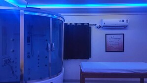 A spa room with a glass-enclosed steam shower, a massage table, and an air conditioning unit on the wall. - Hotel Bramha inn (Pune)