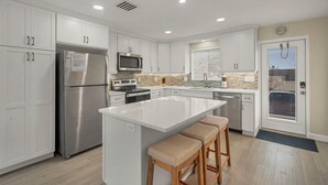 Una cocina moderna con armarios blancos, electrodomésticos de acero inoxidable, una isla central con taburetes y una ventana con persianas.