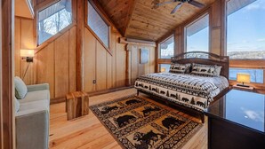 Une chambre dans une cabane en bois, avec un lit, une chaise, un ventilateur de plafond et de grandes fenêtres.