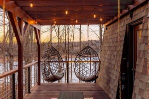 A wooden deck with two hanging chairs, a wooden railing, and a view of a body of water.