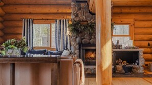L’intérieur chaleureux d’un chalet avec une cheminée en pierre, des murs en bois, un coin repas avec un canapé et une grande fenêtre.