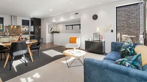 Een moderne woonkamer met een bank, salontafel en een eethoek. In de achtergrond is een keuken met een raam te zien.