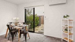 A modern dining area with a table set for four, a white wall, and a sliding glass door leading to a garden.