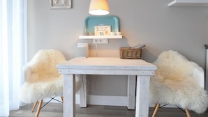 A dining area with a white table, two fluffy chairs, a blue shelf with a sign, and a hanging lamp. - Vacation apartment Bude 16 (Norderney)