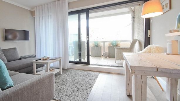 A modern living room with a sofa, a coffee table, a TV, a dining table, and a sliding glass door leading to an outdoor area with plants and a chair. - Vacation apartment Bude 16 (Norderney)