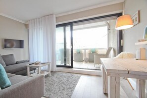 Un salon moderne comprenant un canapé, une table basse, un téléviseur, une table à manger et une porte-fenêtre coulissante donnant sur un espace extérieur agrémenté de plantes et d’un fauteuil.