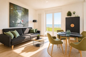 Een moderne woonkamer met een bank, een salontafel en een eettafel met stoelen. Er hangt een schilderij aan de muur, staat een potplant en is er uitzicht op zee.