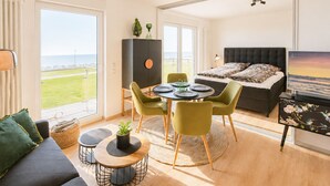 A modern living room with a sofa, dining table, chairs, a cabinet, and a bed with a view of the sea. - Vacation apartment Gezwitschert (Norderney)