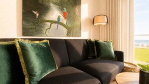 A modern living room with a dark sofa, green pillows, a painting of birds, a lamp, and a view of the outdoors. - Vacation apartment Gezwitschert (Norderney)