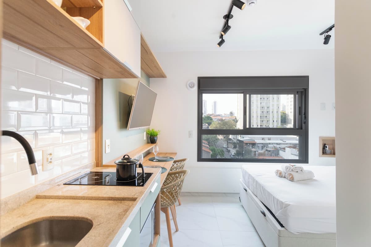 Une cuisine moderne dotée d’une cuisinière intégrée, un coin repas avec une table et des chaises, une grande fenêtre offrant une vue sur la ville, et un lit avec des serviettes soigneusement pliées.