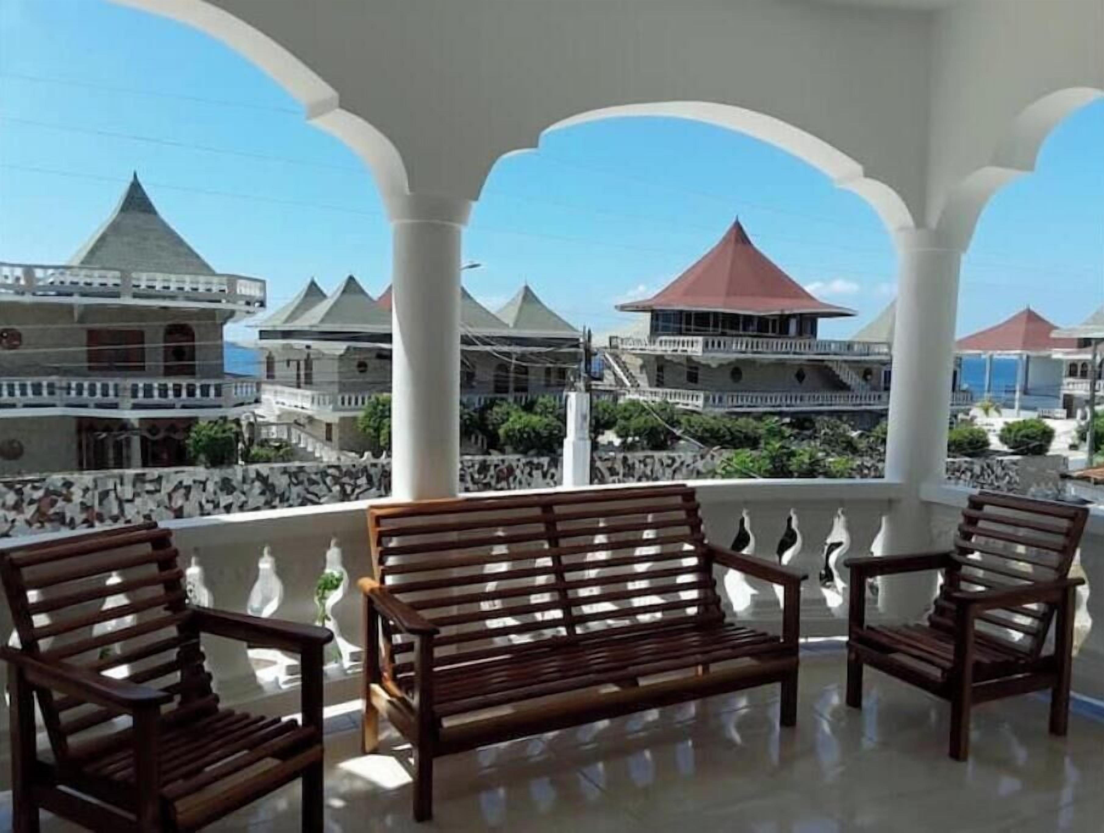 A balcony with wooden chairs and a bench, overlooking a building with multiple towers and a red-roofed structure.