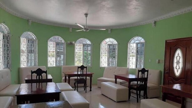 A room with green walls, a ceiling fan, and four windows with decorative grills. There are two sofas, a table, and two chairs.