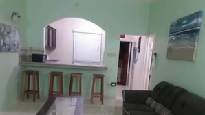 A living room with a dark green sofa, a small wooden table, and a kitchen area with a microwave and a window with blinds.
